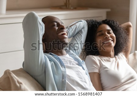 Happy african American husband and wife relax on comfortable couch in living room breathe fresh air, smiling biracial couple rest on cozy sofa have fun enjoying leisure weekend, stress free concept