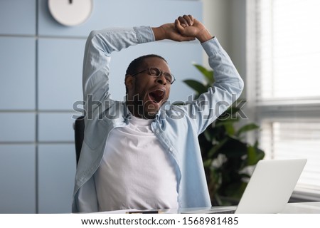 Tired african American male worker yawn stretch in office chair overwhelmed with work at workplace, exhausted biracial young man employee sign feel fatigue after long day, having sleep deprivation Royalty-Free Stock Photo #1568981485
