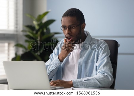 Surprised african American man in glasses work on laptop stunned by unexpected sale offer online, shocked biracial male employee look at computer screen amazed by unbelievable news or notification Royalty-Free Stock Photo #1568981536