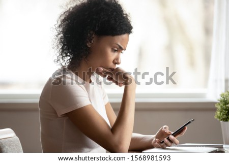 Pensive african American millennial female look at smartphone screen thinking of gadget problem, thoughtful biracial woman using modern cellphone reading news, pondering or considering Royalty-Free Stock Photo #1568981593