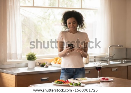 Young african American woman cooking food in modern kitchen using smartphone reading recipe online, millennial biracial female tenant relax at home prepare dinner enjoying free leisure weekend