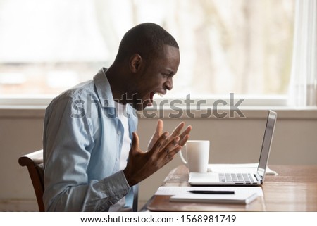 Furious african American male worker look at laptop screen screaming experience operational gadget problems, angry biracial man shout yell feel stressed with computer spam, breakdown or data loss Royalty-Free Stock Photo #1568981749