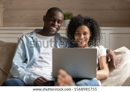Smiling millennial african American couple relax on comfortable couch in living room watching video on laptop, happy biracial husband and wife rest on cozy sofa browsing web shopping online together