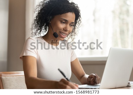 Focused african American female student in earphones study watch webinar or distant course on laptop making notes, concentrated biracial woman in headphones work on computer consult client online Royalty-Free Stock Photo #1568981773
