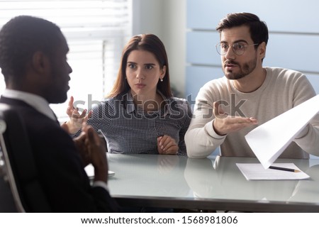 Angry Caucasian millennial couple customers dispute argue with african American realtor or broker show error in documents, mad frustrated clients blame biracial male agent for mistake, money fraud Royalty-Free Stock Photo #1568981806