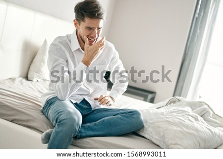 Jeans a white shirt A young man is sitting on a bed and a window is in the background