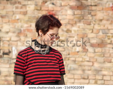 Alternative girl in pink sunglasses on a brick wall background with copy space. Goth girl with a short haircut and neck collar looking down