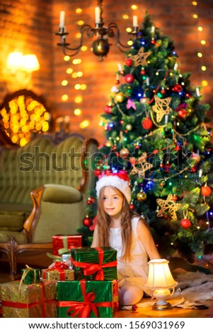 A cute girl in a white dress and Santa Claus hat is sitting on the floor with a large pile of gifts. Against the background of a festive tree. Christmas and New Year