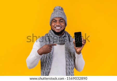 Smiling african american winter man holding latest slim smartphone with blank screen and point at it. Trendy mobile phone, copy space