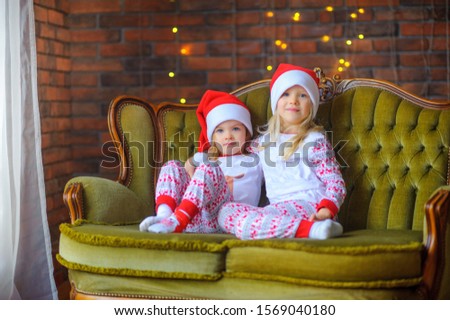 Two cute little sisters in a santa hat are sitting on the sofa in the room. Waiting for the holiday
