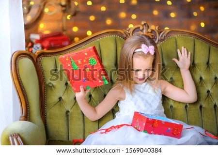 Happy little girl in a dress sits on a sofa with a gift box in her hands opens and smiles. Christmas and New Year