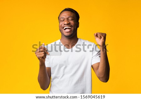 Mobile Games. Happy African American Man Holding Cellphone Shaking Fists Standing In Studio Over Yellow Background.