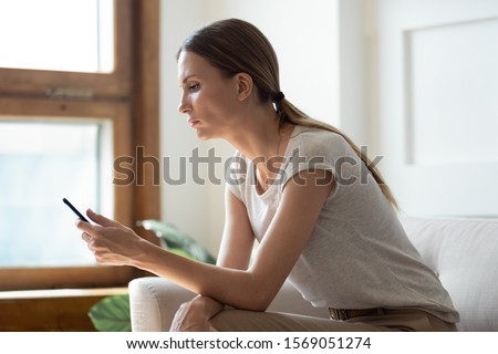 Side view thoughtful young woman sitting on couch, looking at mobile phone screen, waiting for message from beloved man, feeling lonely or jealous. Unhappy lady reading news in social media.