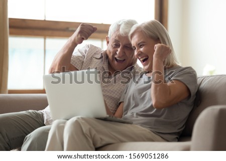 Euphoric older married spouse looking at computer screen, making yes gesture, celebrating online lottery win or cheering favorite sport team. Overjoyed retired couple received email with good news. Royalty-Free Stock Photo #1569051286
