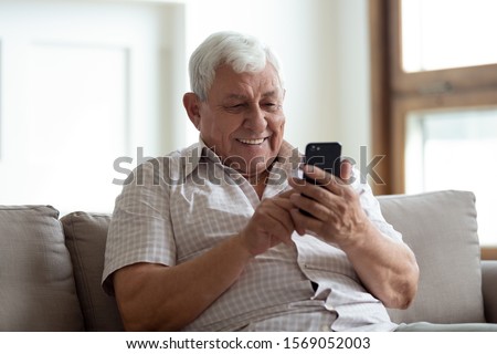 Happy older man sitting on comfortable sofa at home, using smartphone. Pleasant 80s grandfather reading sms with good news, typing message for grown up kids, shopping in mobile app, watching photos. Royalty-Free Stock Photo #1569052003