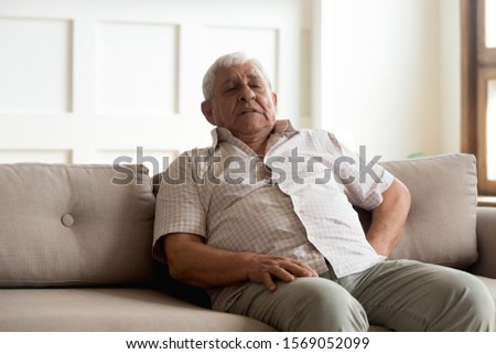 Unhealthy older man leaning on couch, suffering from strong pain in back. Unhappy elder grandfather feeling unwell, waiting for ambulance aid, resting alone on sofa at home, reducing painful feelings. Royalty-Free Stock Photo #1569052099