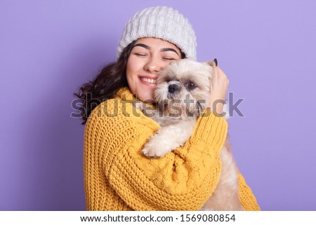 Indoor picture of young beautiful curly haired lady holding her Maltese dog in hands, closing eyes, having pleasant facial expression, closing eyes, pressing it close to face. People and pet cocnept.
