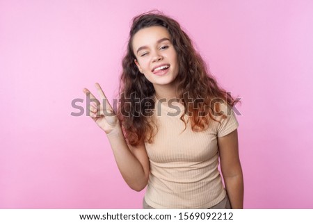 Portrait of cheerful vivid teenage girl with long curly brunette hair wearing casual style beige clothes winking at camera and showing victory gesture. indoor studio shot isolated on pink background