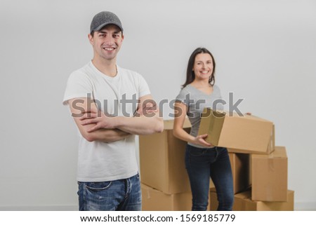 Strong delivery man transported boxes into new apartment of lovely young woman and is standing on the forefront, his hands folded. Pleased woman is standing on the background, blurred, holding a box.
