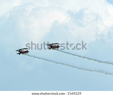 Color DSLR picture of two stunt planes at an airshow.  Biplanes are speeding horizontally, trailing white smoke contrails against cloudy sky. Airplanes are dangerous and fast.  Copy space for text.