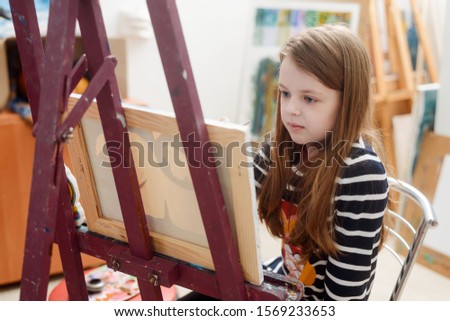 Little girl learning to draw, sitting on a chair in front of a canvas. Beginner student in fine art class.