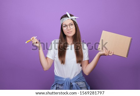 A beautiful girl with bright emotions, with dark hair, in a white T-shirt and bandana, is holding a brown notebook and pen in her hands. Makes a choice. Photo on a purple background.