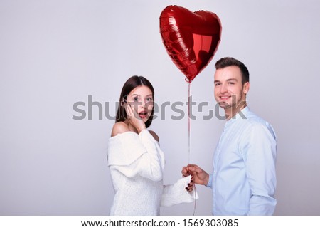The young brown-haired woman opened her mouth in surprise, receiving a gift from a red inflatable heart. A man in a blue shirt gives his sweetheart a red heart-shaped balloon. Lovers on background.