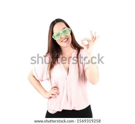 Happy young woman doing an ok gesture with her hand while wearing green sunglasses against a white background
