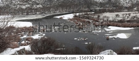 Snow fell on dry yellow grass on banks of river. Bank of river after first snowfall. River flows along hilly snow-covered shore. Beautiful winter landscape. Cloudy winter day. Horizontal photo.