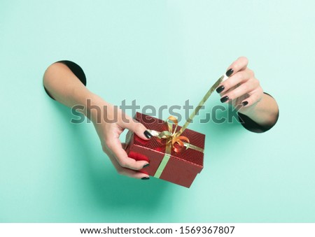 Female hands hold red gift through a hole on neon mint background.