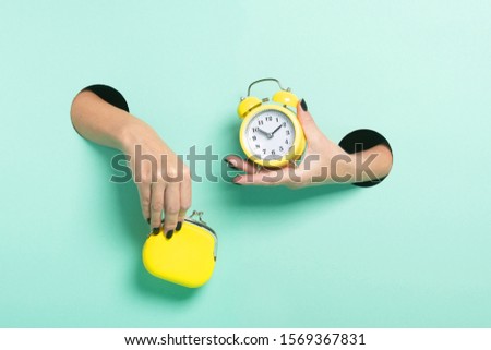 Female hands hold alarm clock and wallet through a hole on neon mint background. Time is money concept.