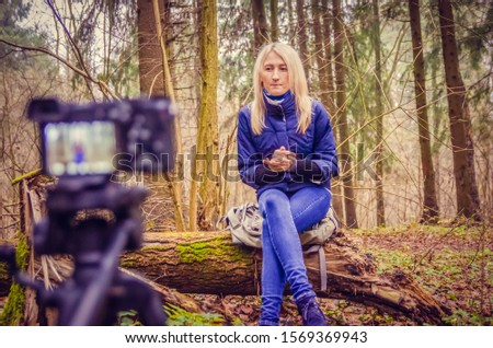 blonde girl in a blue jacket and jeans, boots writes a blog like a blogger sitting in a forest, Park on a fallen tree. Writes a blog on the DSLR.