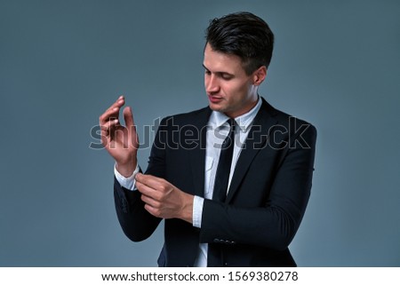 Portrait of trendy, attractive, stunning man in black tuxedo with tie fasten button on sleeve cuffs of his white shirt, isolated on grey background.