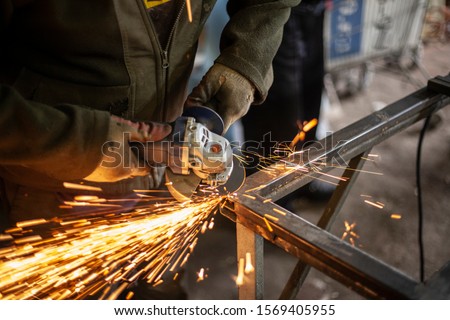 A man polishes metal in a workshop. Creation of an advertising structure. Metal frame in the garage. Sparks from metal cutting. Vivid flashes he split parts and due to friction. Royalty-Free Stock Photo #1569405955