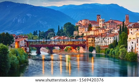 Bridge Ponte degli Alpini at river Brenta Bassano del grappa Italy. Panoramic view at old town with vintage building and tower and wooden bridge at background Alpine mountains scenic landscape. Royalty-Free Stock Photo #1569420376