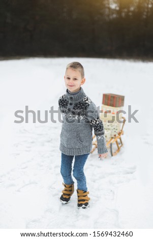 Little boy with Christmas present boxes on wooden sled in snow. Little boy with sledge in snowy forest. Winter holidays decoration. Child with sleigh. Kid sledding.
