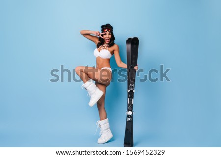 Emotional woman in bikini and goggles dancing on blue background. Studio shot of laughing girl in white swimsuit standing on one leg and holding skis.