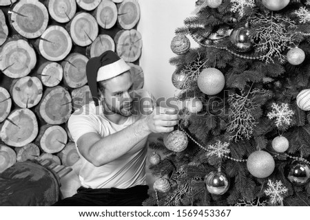 Man in santa claus hat decorate Christmas tree with balls, snowflakes, garlands on wood logs background. xmas, new year, eve, holidays celebration. Festive decorations and ornaments
