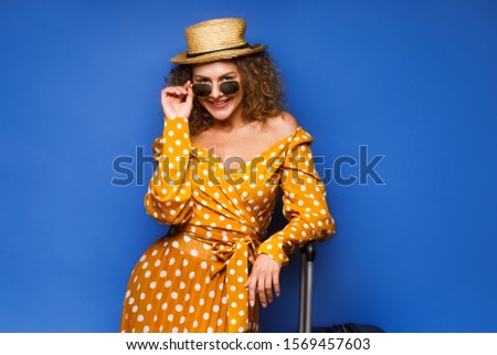Young woman in vintage dress stay on background holding suitcase. Amazed curly european girl posing before vacation with her luggage. Traveling time.