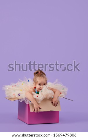 A cheerful little girl child in a magnificent holiday dress sits inside a large gift colored box, on a purple isolated background. Birthday party surprise. Space for text