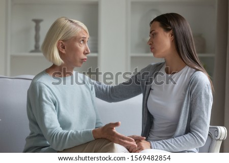 Grown up daughter hold hand of 50s elderly mother, women seated on couch in living room having talk, mom pout lips looks helpless receiving support from adult child in difficult period of life concept Royalty-Free Stock Photo #1569482854