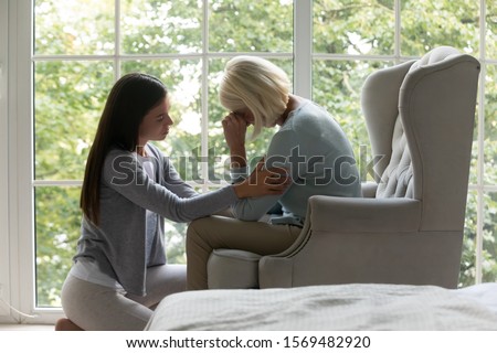 Near panoramic window crying elderly mom sit on armchair, near sit her grown up daughter comforting her in difficult life period give support, share pain at divorce, showing attention and care concept Royalty-Free Stock Photo #1569482920