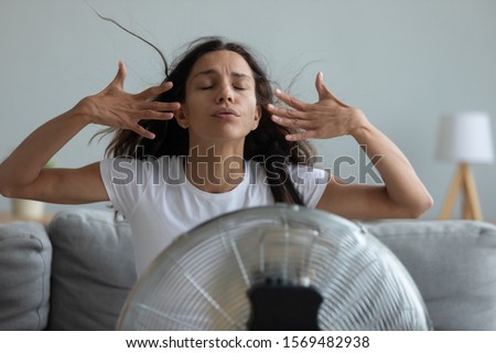 Woman seated on couch in living room suffers from swelter waving her hands trying to cool off by the fan, oppressive heat too hot weather and sultriness in summer, flat without air-conditioner concept Royalty-Free Stock Photo #1569482938