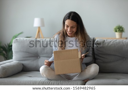 Overjoyed woman seated on sofa hold on lap small cardboard box open parcel client feels satisfied bought goods in internet, happy addressee received package from friend, quick delivery service concept Royalty-Free Stock Photo #1569482965