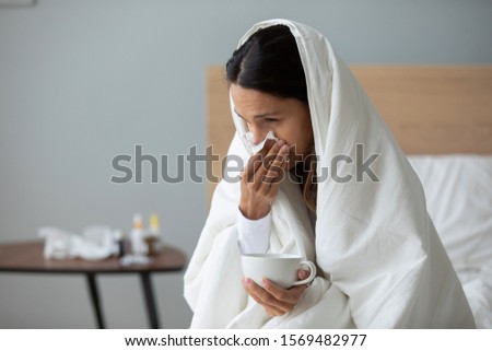 Sick woman froze and caught a cold from an unheated flat covered with warm blanket sit in bed drinking cup with hot beverage or medicines using handkerchief blows runny nose, seasonal grippe concept Royalty-Free Stock Photo #1569482977
