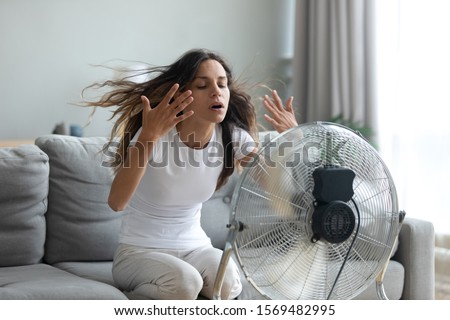 In living room without air-conditioner tired from summer heat young woman turned on floor ventilator waving her hands to cool herself, female sitting on couch suffers from unbearable too hot weather Royalty-Free Stock Photo #1569482995