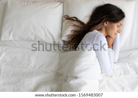Top above view single serene young woman wearing white sleepwear sleeping lying on one side of bed in the bedroom on comfortable pillow orthopaedic mattress enjoy sweet dreams healthy posture concept Royalty-Free Stock Photo #1569483007
