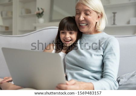 Granddaughter and grandma activity with modern technology concept, cheerful seven years old girl leaned on couch sit together with grandmother using laptop, watching cartoons, enjoy distant video call