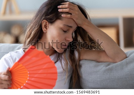 Close up view millennial woman sitting on couch exhausted by hot weather closed eyes feels unwell dying of heat holding in hand orange fan using it for reducing too hot temperature indoors concept Royalty-Free Stock Photo #1569483691