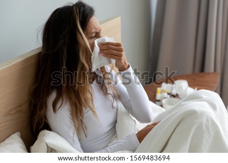 Sick 30s biracial woman sitting on bed, covered with white blanket sneezing holding paper handkerchief, wiping blowing runny nose, feels unhealthy suffering from seasonal grippe, caught a cold concept Royalty-Free Stock Photo #1569483694